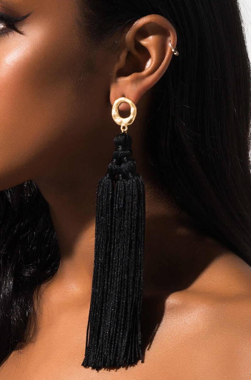Close-up of a person wearing the ON THE FRINGE EARRING, featuring intricate black tassels hanging from a gold hoop at the top. The earrings are long and dangle down from the earlobe. The person has smooth, dark skin and straight, dark hair with no other facial features visible.