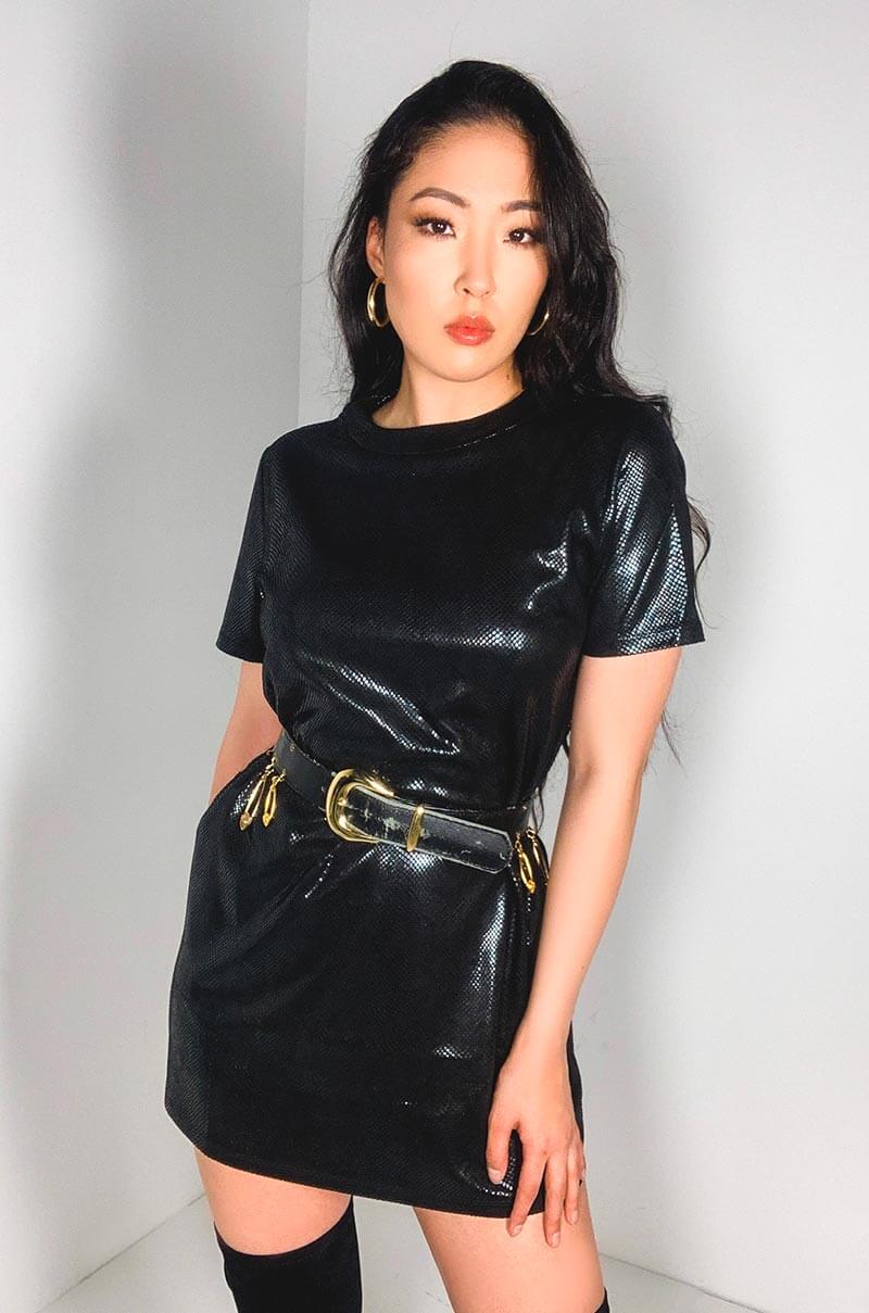 A woman with long dark hair stands against a plain white background, dressed in the ONLY THING I GOT MINI DRESS made of shiny black material. She accessorizes with a black and gold belt, gold hoop earrings, and knee-high black boots.