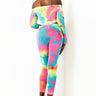 A person stands against a white background, facing sideways and looking back. They are wearing a vibrant, colorful off-the-shoulder bodysuit that features an eye-catching tie-dye pattern in yellows, greens, blues, and pinks. They also sport the clear ITS MY TIME MESH LEGGING along with clear high heels.