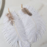 Product Description: The PAULA FEATHER EARRING is displayed on a light marble background, featuring white feathers with intricate brown and black details near the top. One feather displays a delicate polka-dotted pattern. The earrings are completed with elegant gold hooks.