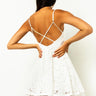 A woman with long dark hair stands with her back to the camera, showcasing the elegant back design of the PARTY PLAY LACE DRESS. The sleeveless dress features thin straps that cross at the back and intricate white lace fabric. With her hands resting on her hips, she highlights its stunning details.