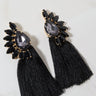 Introducing the LUXY FRINGE EARRING, a pair of sophisticated black tassel earrings. Each earring showcases a teardrop-shaped black gemstone encircled by smaller black stones, beautifully arranged in a fan-like pattern. Elegant long black tassels dangle gracefully below the gemstone cluster, making these earrings perfect for any occasion. Photographed on a light surface, they're sure to catch your eye.