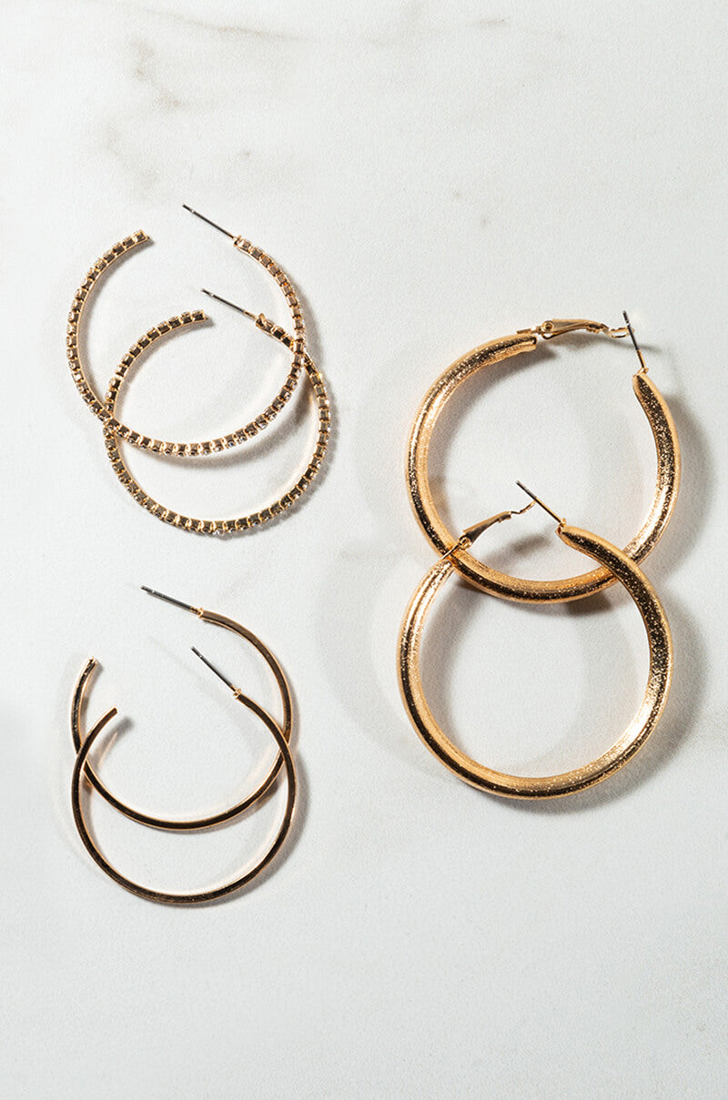 Displayed against a light surface, the "PERFECT PAIR HOOP SET" showcases four pairs of gold-tone hoop earrings. From left to right: a small pair adorned with rhinestones, a larger textured pair, a medium-sized smooth pair, and another small pair featuring a double hoop design.