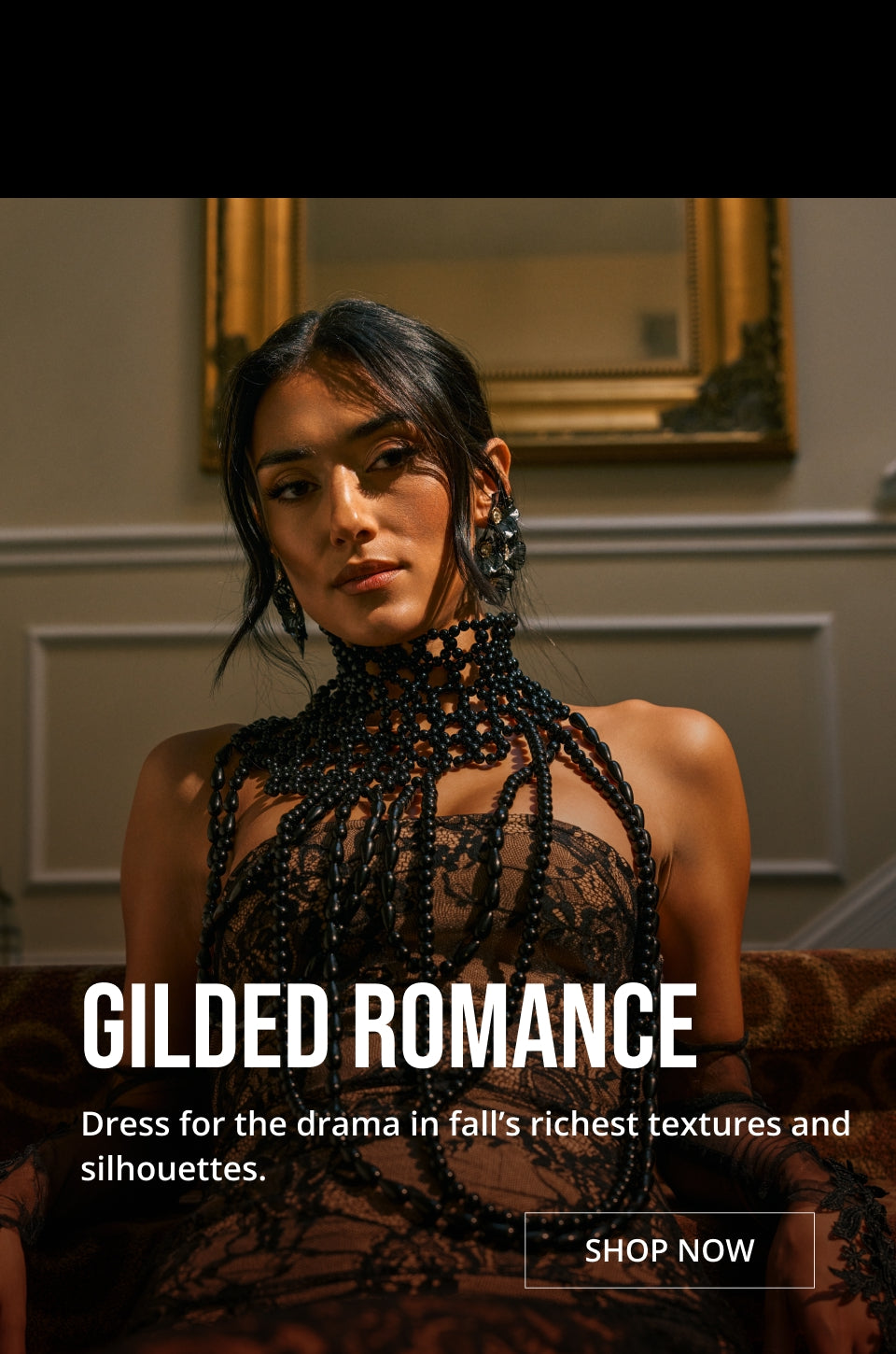 A woman wearing a black lace and beaded high-neck dress with sheer draped details, posed indoors in front of a mirror. Large text reads 'GILDED ROMANCE' with smaller descriptive copy and a 'SHOP NOW' button.
