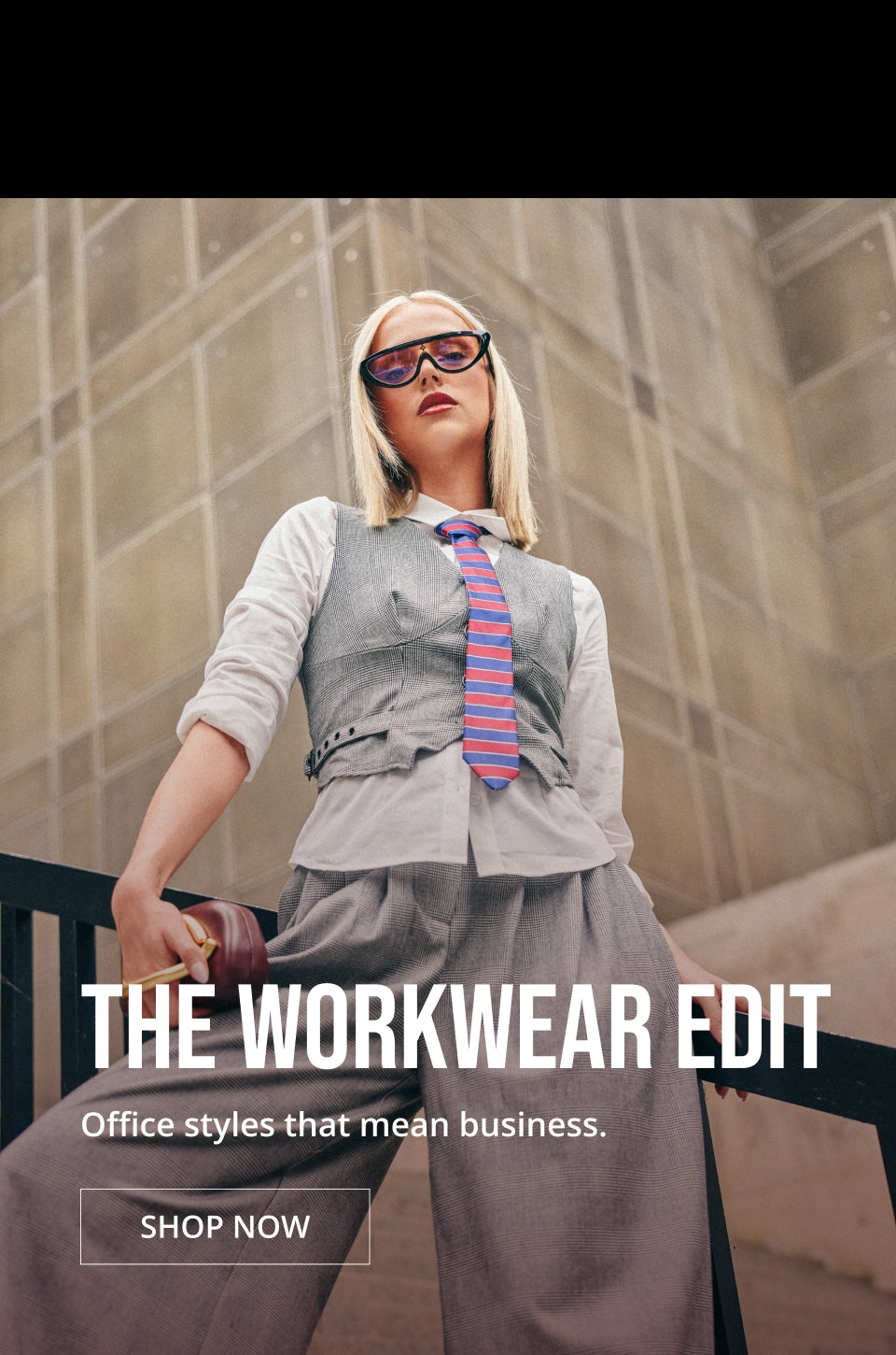 A fashion editorial-style photo of a woman wearing a gray vest, white shirt, striped tie, and sunglasses, posed against a stone building. Large overlaid text reads 'THE WORKWEAR EDIT' with the subtitle 'Office styles that mean business.