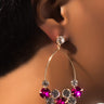 Close-up of a person wearing the POURING DANGLE EARRING by Mika Accessories. The gold earring has a circular cluster design adorned with sparkling gemstones in pink, green, and clear. The person has dark hair and a medium skin tone.