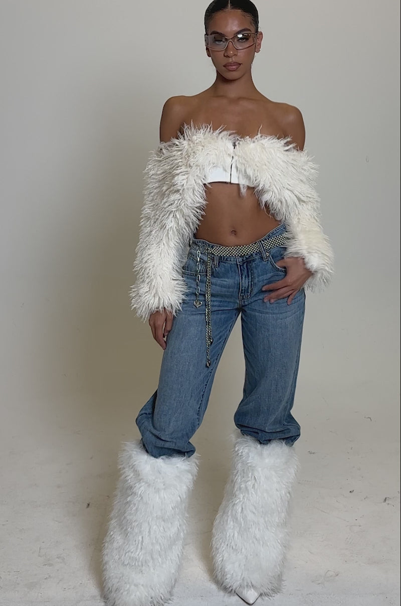 A video of a woman in the SNOW FUR TOP off-the-shoulder, paired with matching furry leg warmers, blue jeans, a chain belt, and sunglasses against a plain light background.