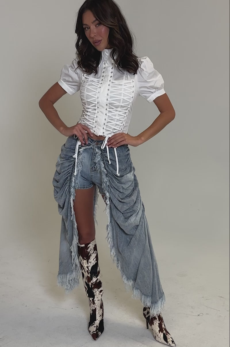 Video of woman modeling the ALL OVER THE PLACE DENIM SHORT with distressed detailing and fringed panels, and knee-high cow-print boots against a plain background.