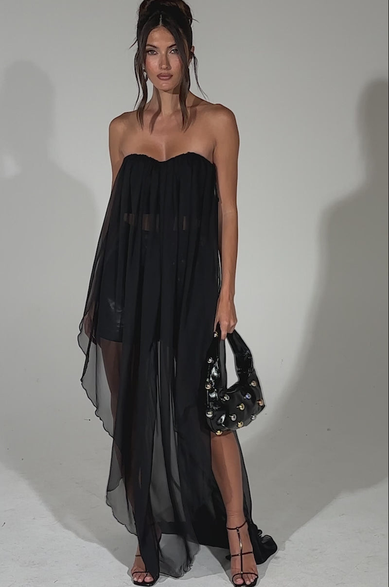 A video of a woman modeling the GONE TOO LONG BLOUSE, a black sheer top with a sweetheart neckline and chiffon overlay featuring a long, flowing train over black shorts. She accessorizes with a black handbag, strappy heels, and wears her hair in a high ponytail.