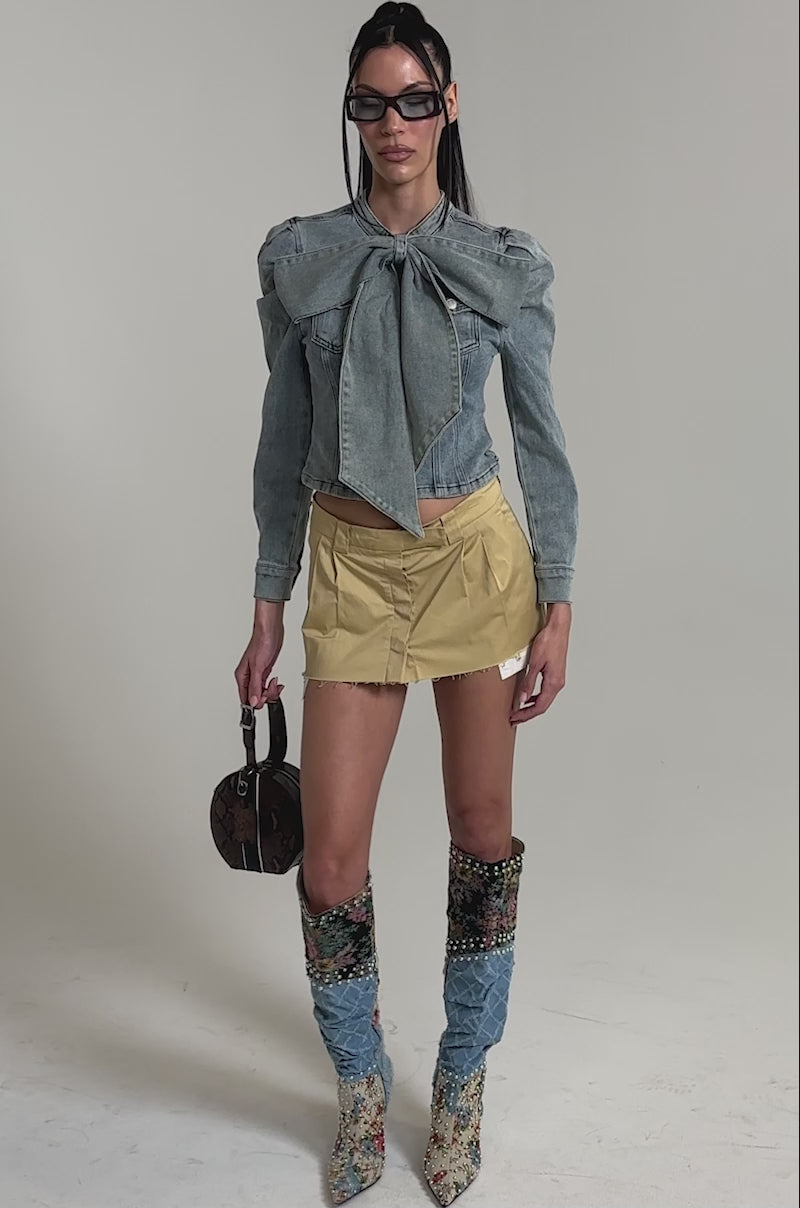 A video of a woman wearing the WAIT FOR U DENIM BOW BLOUSE TOP with puffed shoulders, a short beige skirt, floral and denim knee-high boots, sunglasses, and holding a round brown handbag.