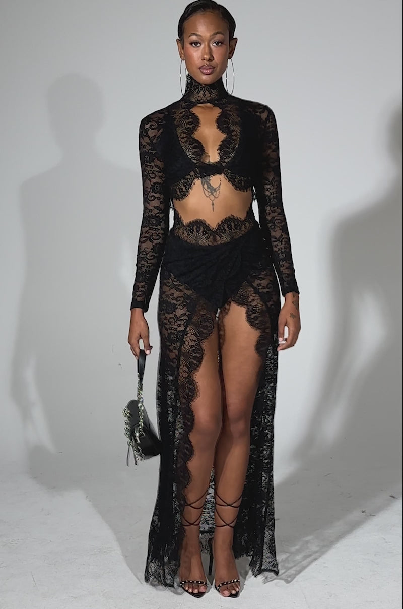 A video of a woman posing in the LAY IT LOW LACE MAXI DRESS, featuring sheer floral lace, long sleeves, and a high slit skirt. She accessorizes with a black handbag with silver rings, strappy black heels, and hoop earrings.