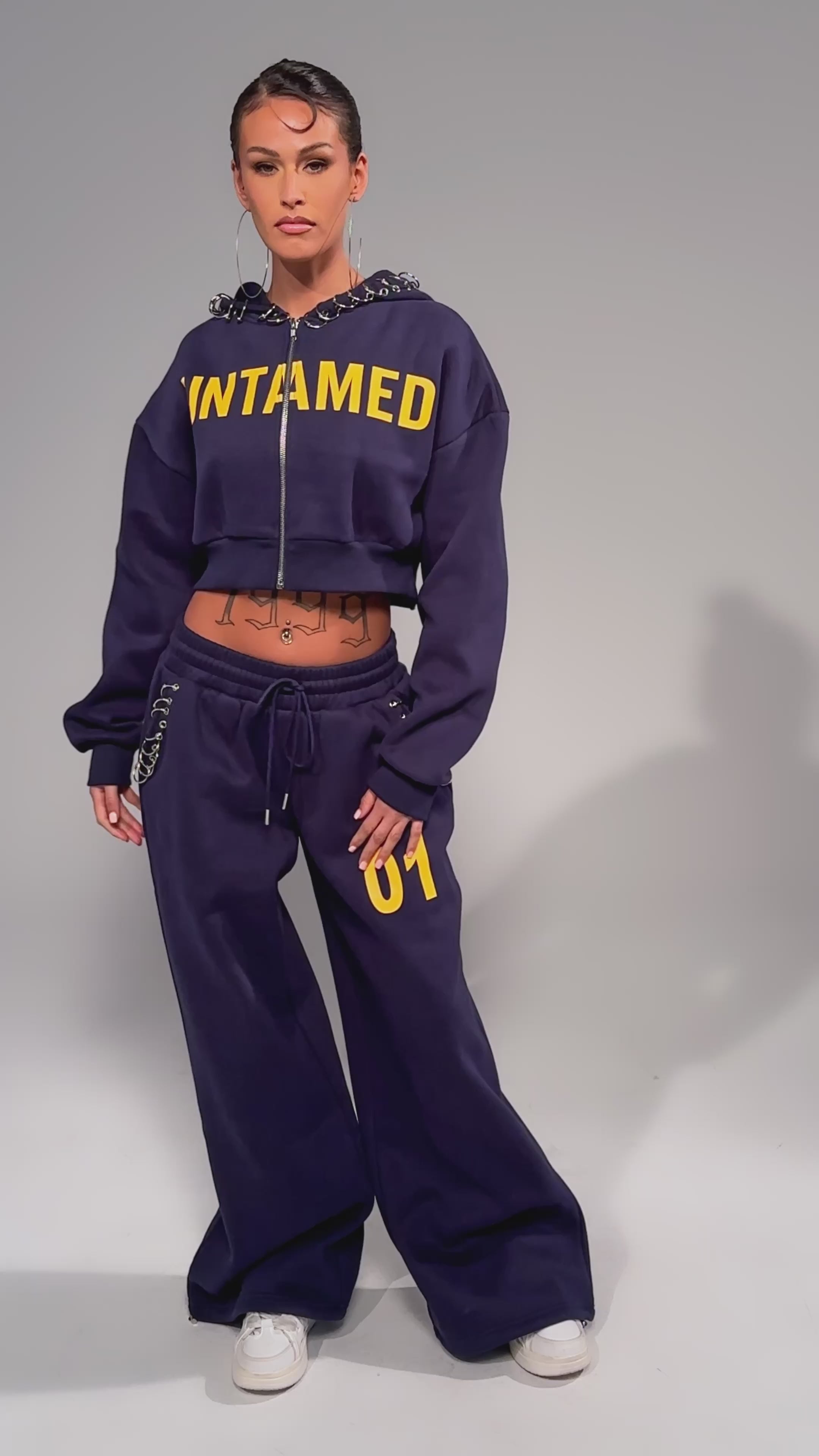 Video of woman modeling the UNTAMED METAL RING JOGGERS—navy, wide-leg, high-rise with "01" on one leg—paired with a matching cropped hoodie featuring "UNTAMED" in yellow, hoop earrings, white sneakers, and visible tattoos.