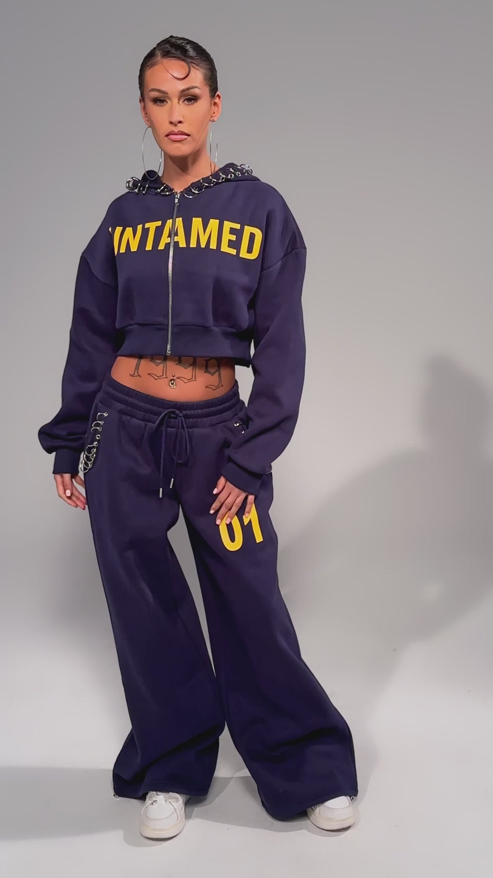 Video of woman modeling the UNTAMED METAL RING JOGGERS—navy, wide-leg, high-rise with "01" on one leg—paired with a matching cropped hoodie featuring "UNTAMED" in yellow, hoop earrings, white sneakers, and visible tattoos.