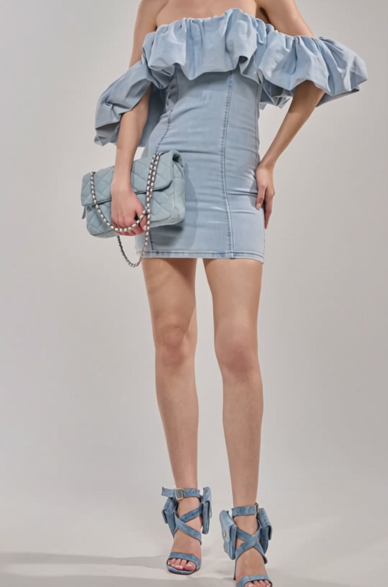 Video of woman modeling the JULIA DENIM MINI DRESS, a light blue off-the-shoulder mini dress with ruffles. She accessorizes with a quilted chain-strap purse, gold hoop earrings, and wears her long hair down.