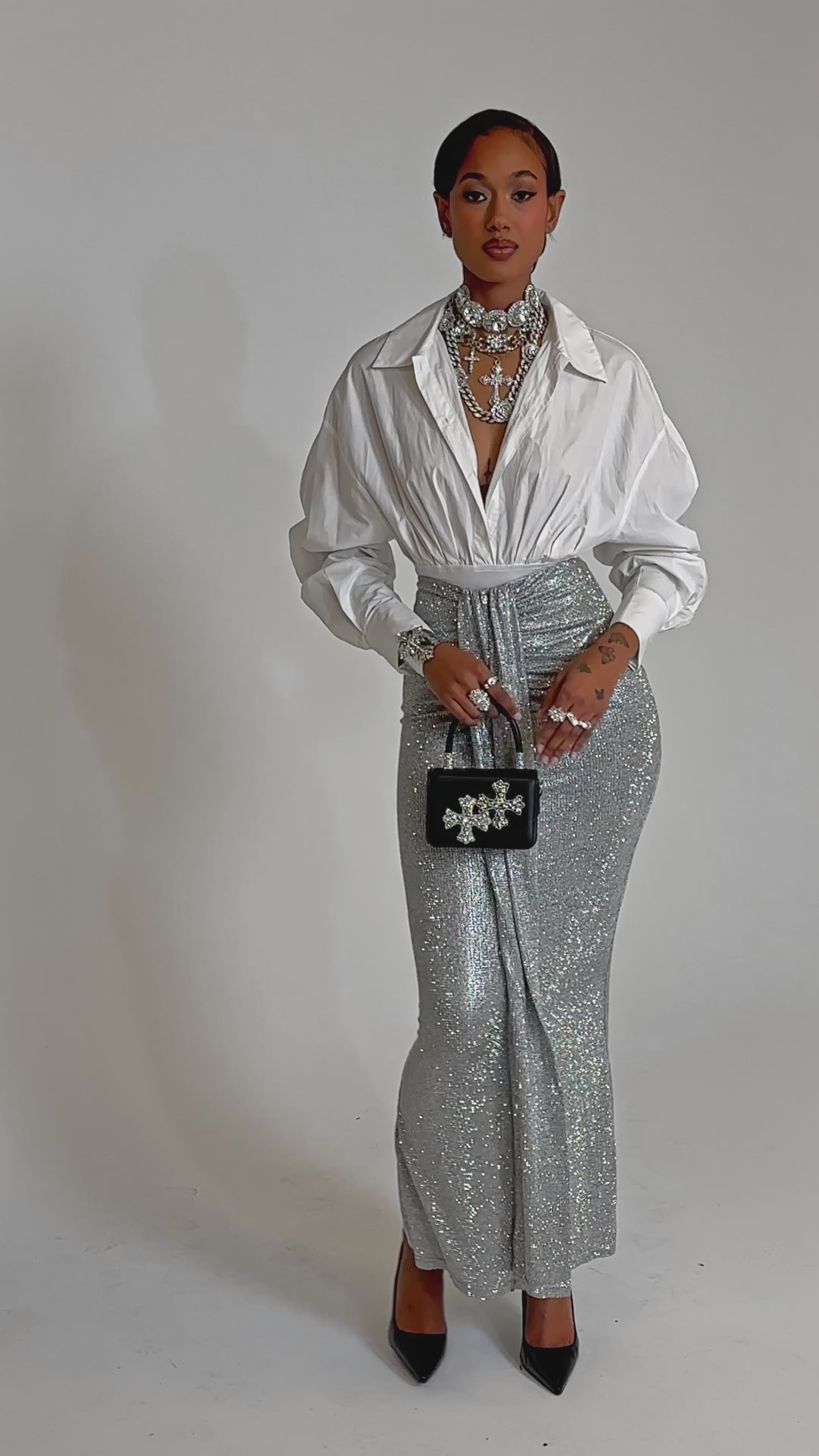 A video of a woman wearing the BRIGHT LIGHTS RHINESTONE MAXI SKIRT IN SILVER, a white deep-neck blouse, black heels, layered silver jewelry, and holding a small black handbag—an ideal chic look for evening events.