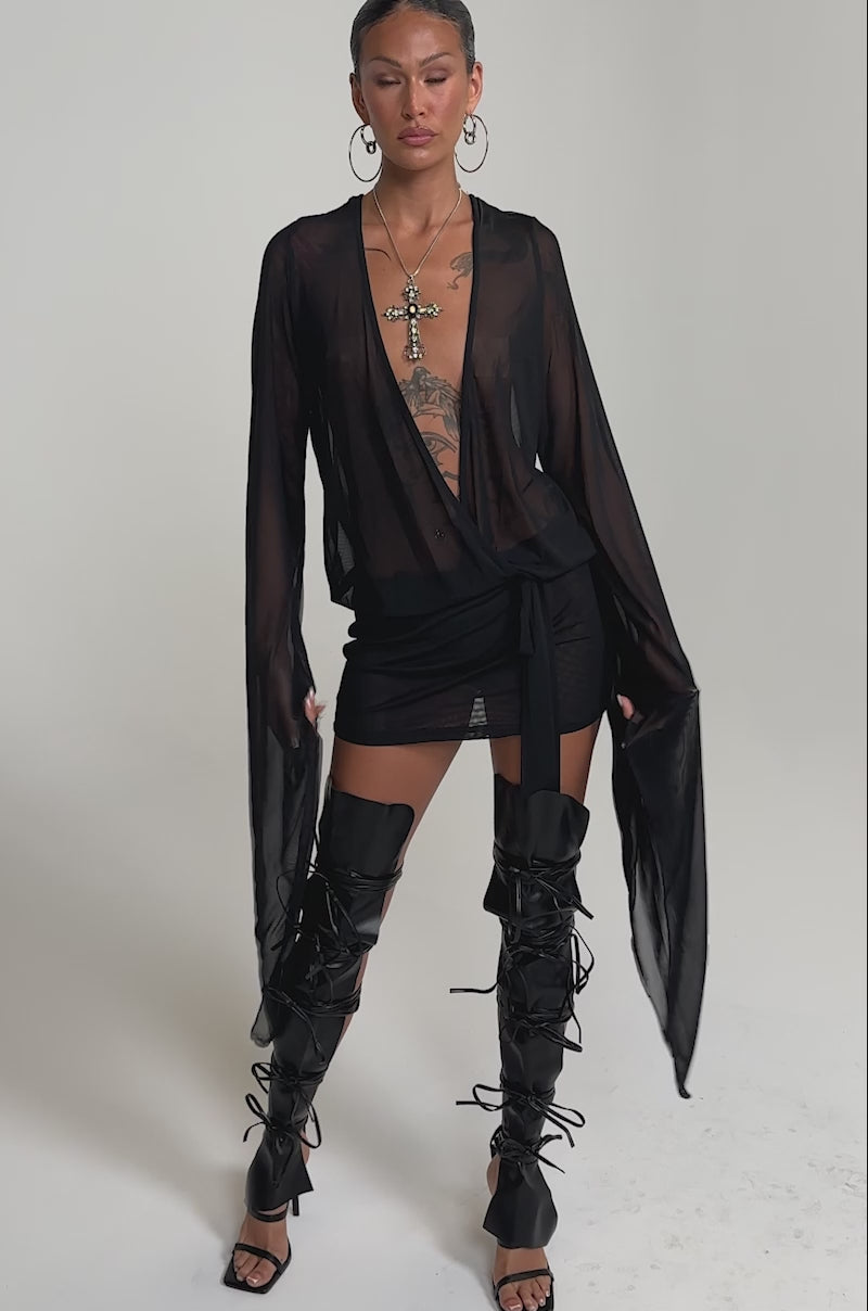 A video of a woman wearing the SHEER AFFECTION MESH MINI DRESS, styled with thigh-high lace-up boots, large hoop earrings, and layered necklaces.