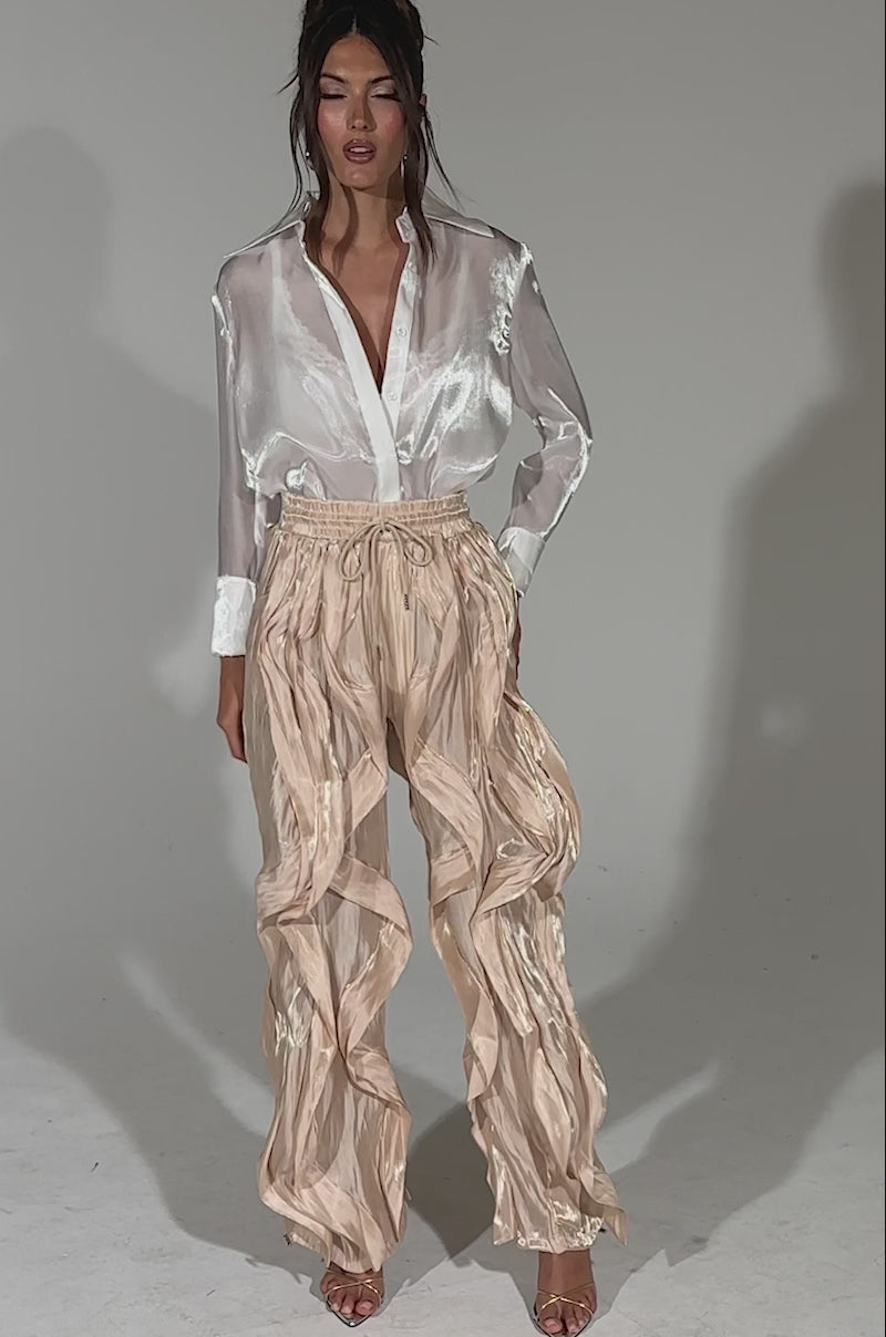 A video of a woman standing against a plain light background wearing the COOLEST ON THE BLOCK SATIN JOGGER, styled with a shiny white blouse, hands in pockets, showcasing the jogger’s flowing wide-leg fit and ruffle details.