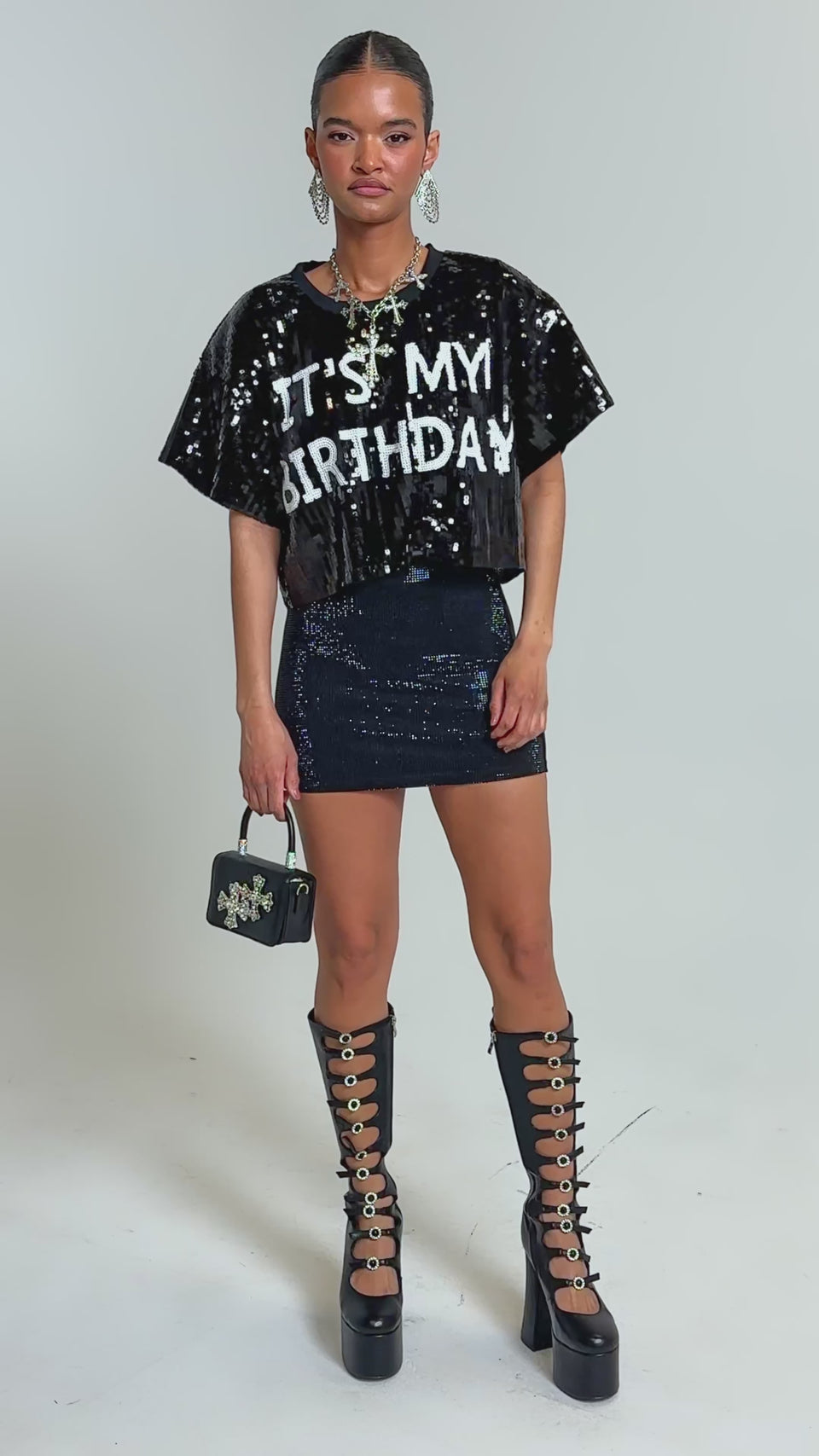 Video of woman modeling the IT'S MY BIRTHDAY SEQUIN T-SHIRT, a black sequin skirt, statement earrings, and a small black handbag with a jeweled cross design