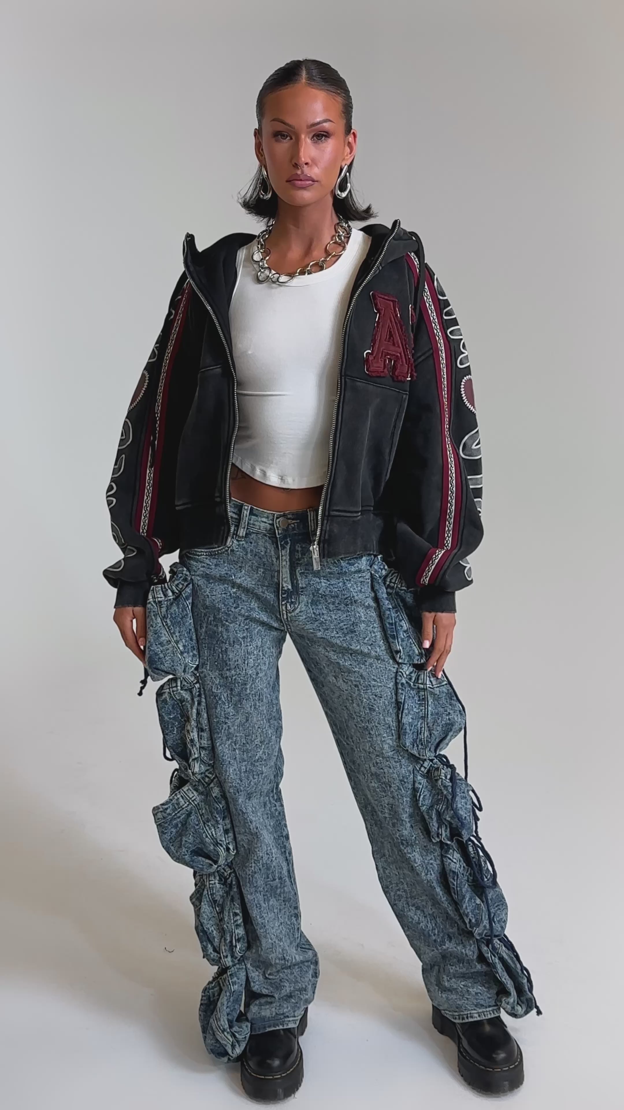 A video of a woman wearing a black zip-up hoodie with bold white sleeve text, a white crop top, POCKETS FULL OF SUNSHINE DENIM PANT featuring high-rise acid-wash and large ruffled sides, chunky black shoes, and a statement necklace.