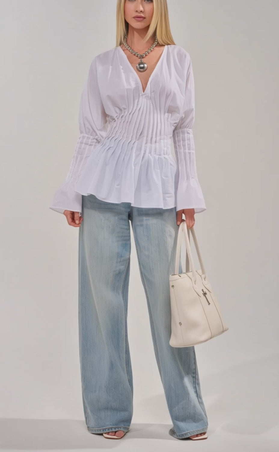 Video of woman modeling the DONT LOOK AT ME BLOUSE, a pleated white top with flared cuffs, paired with light blue jeans and a chunky silver necklace while holding a white bag against a plain light background.