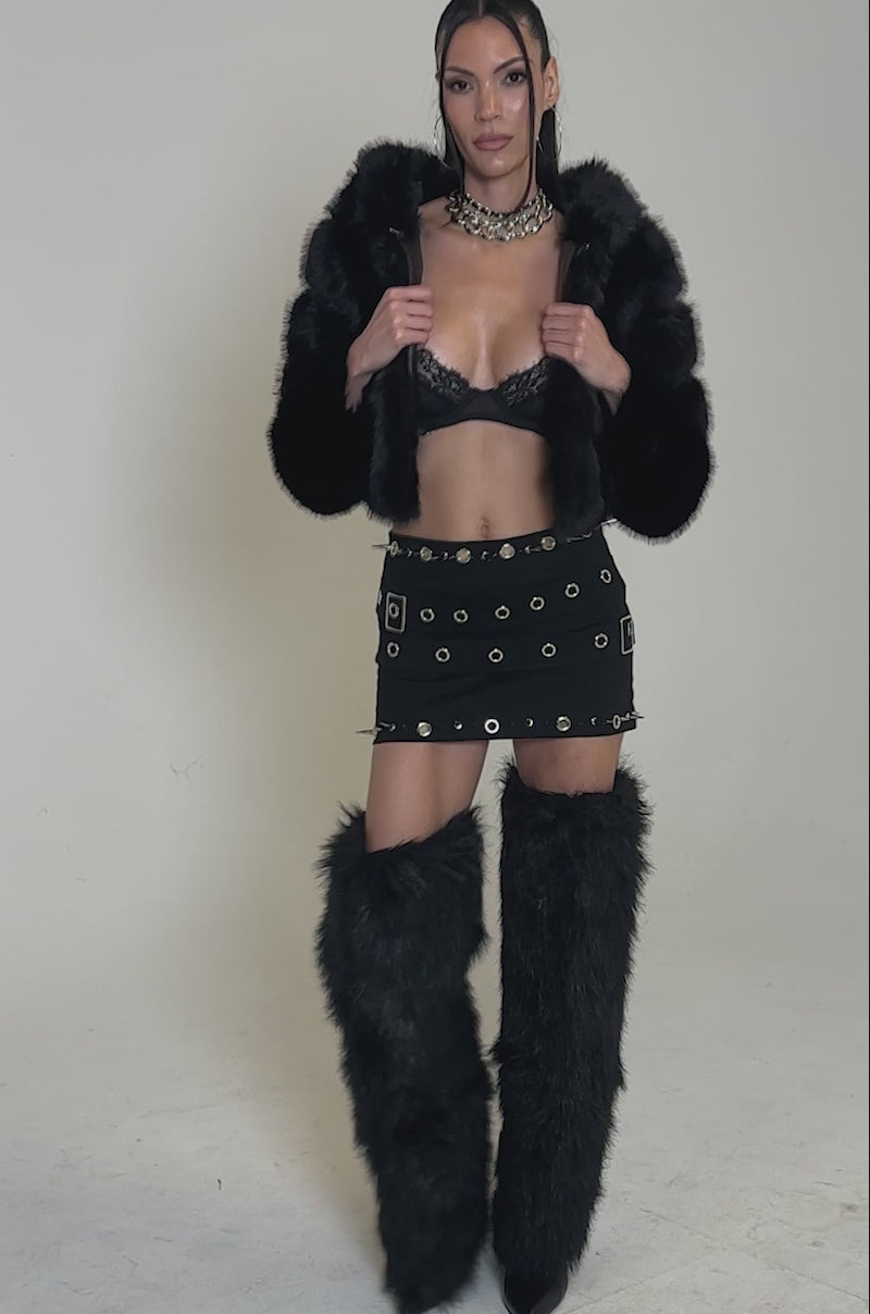 A video of a woman posing confidently in the JOLENE FAUX FUR COAT IN BLACK, paired with a black bra, studded mini skirt, tall black furry leg warmers, layered necklaces, and long straight hair styled in two small top knots.