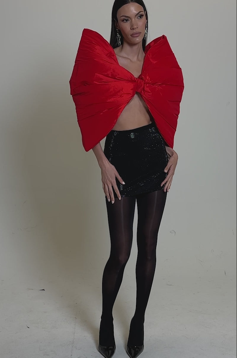A video of a woman wearing a bold red oversized bow-shaped top, the TOO HOT FOR YOU RHINESTONE MINI SKIRT in black, sheer black tights, and black pointed-toe heels.