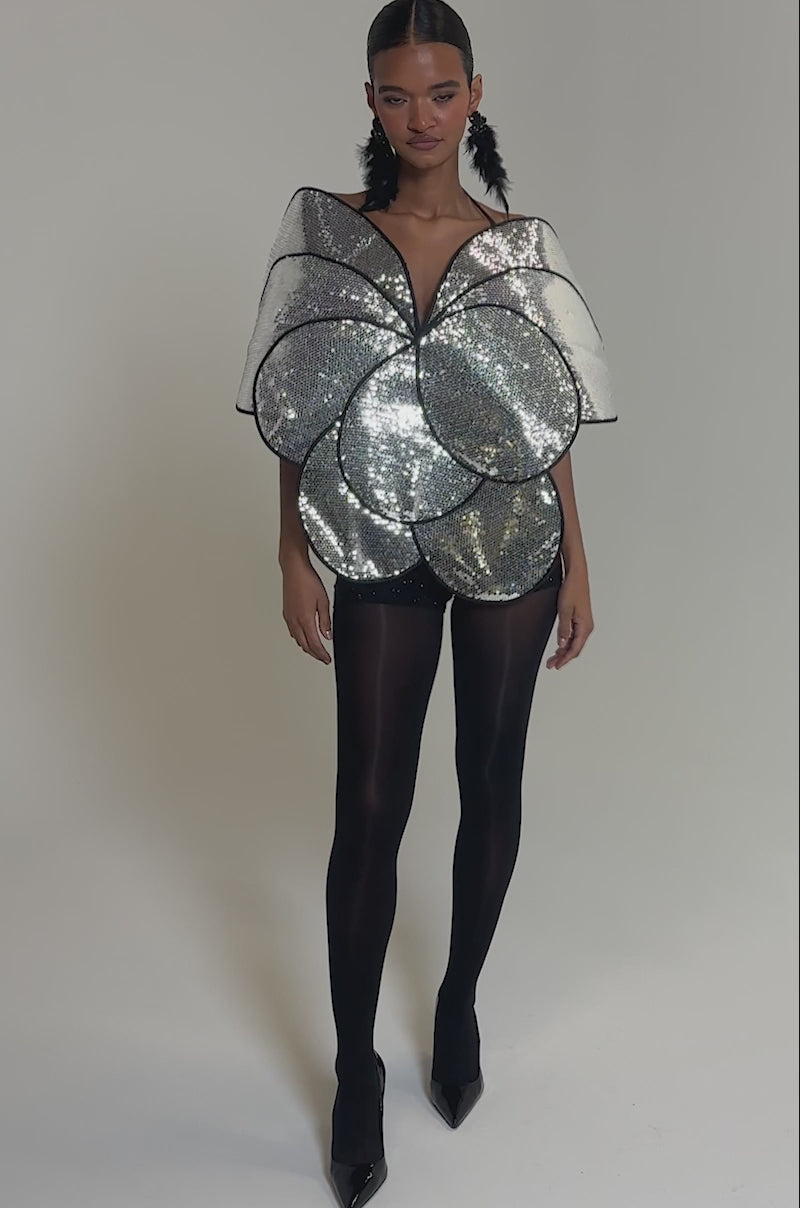 Video of woman modeling the LA DIVINA SEQUIN TOP—an oversized, silver, flower petal-shaped top with sequins—paired with black shorts and sheer tights against a plain gray background.