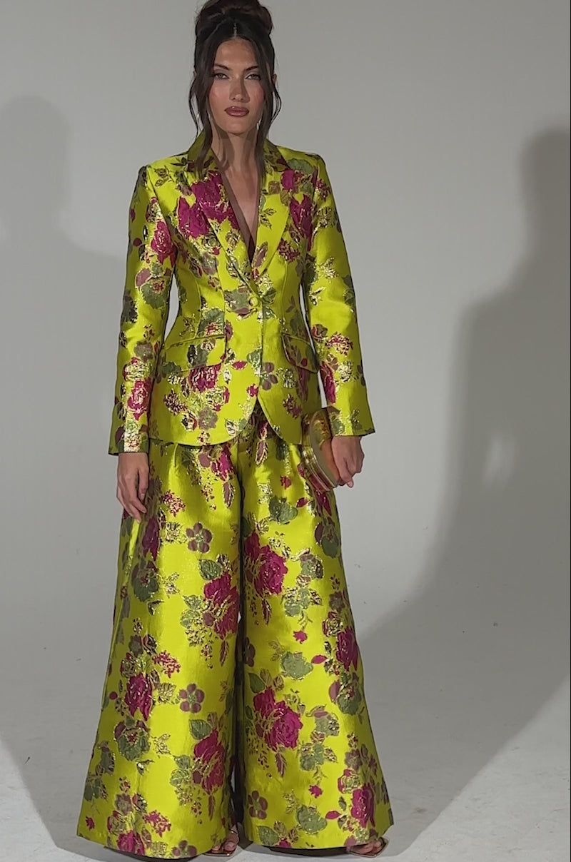 A video of a woman wearing the BUY MYSELF FLOWERS BROCADE TROUSER IN LIME, featuring a shiny lime green hue and pink-gold floral pattern, paired with a matching fitted jacket. Her hair is styled in a loose updo against a plain light background.