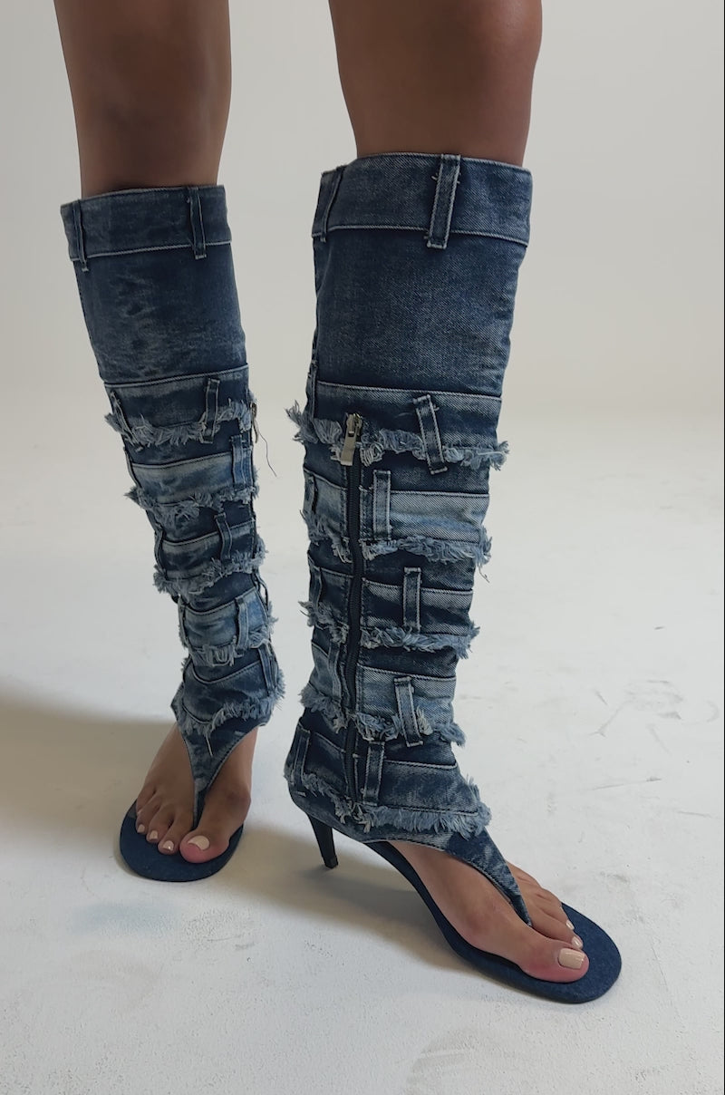 A video of a person standing on a light surface wearing a blue denim mini skirt and the AKIRA GOLD MAHONEY DENIM PANT DETAIL GLADIATOR STILETTO, featuring frayed edges, zippers, and distressed denim heels.
