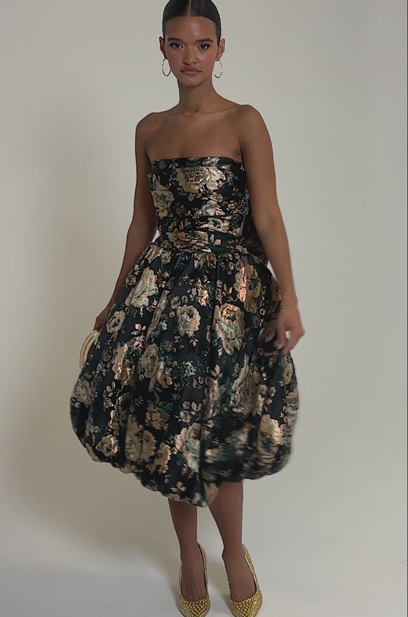 Video of woman modeling the DRESSED TO IMPRESS BROCADE MIDI DRESS, featuring a strapless bodice and voluminous skirt. She completes her look with gold hoop earrings, textured heels, and slicked-back hair.