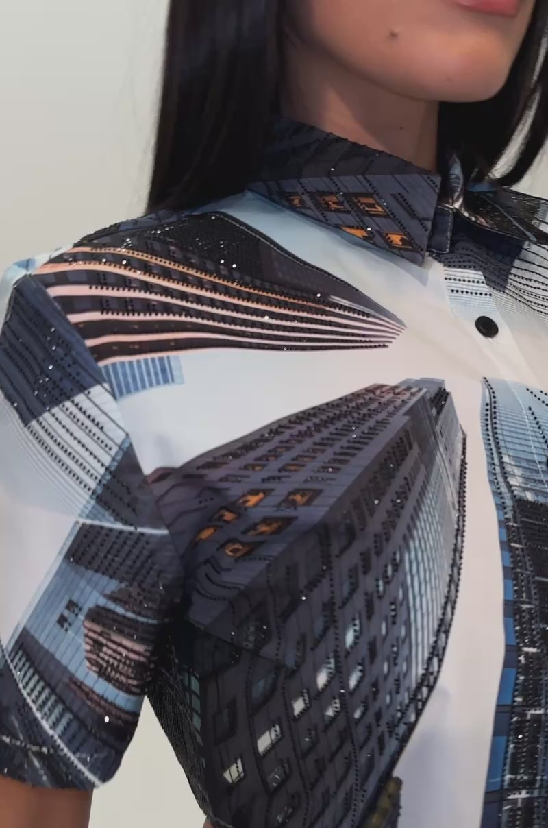 A video of a woman wearing the CITY DREAMS POPLIN BLOUSE—a cropped button-up shirt adorned with vivid skyscraper cityscape prints—and pairs it with a light blue denim skirt. 
