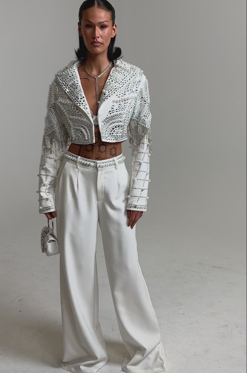 A video of a woman in the DIRTY MARTINI EMBELLISHED TROUSER IN WHITE, paired with a cropped embellished jacket, sheer top, silver heels, and a small white handbag.