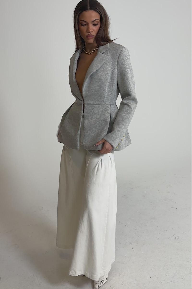 Video of woman modeling the CANARY PEPLUM BLAZER IN SCUBA in light gray, featuring a deep V-neck and sculptural peplum, styled without an undershirt, high-waisted white pants, and a chunky gold necklace against a plain background.