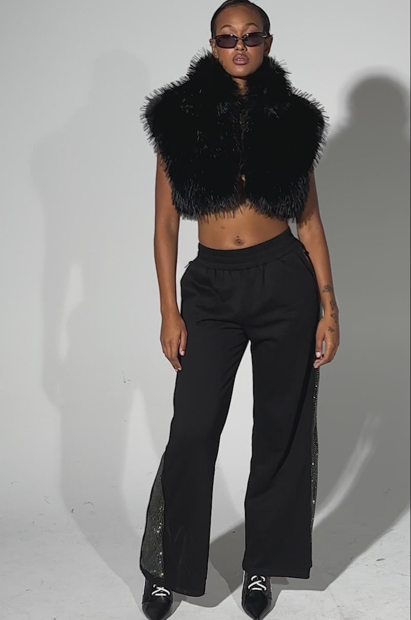 A video of a person wearing black sunglasses, a sleeveless black faux fur vest, the OPEN UP WIDE LEG JOGGER WITH RHINESTONE, and black pointed-heeled boots stands confidently against a plain light background.
