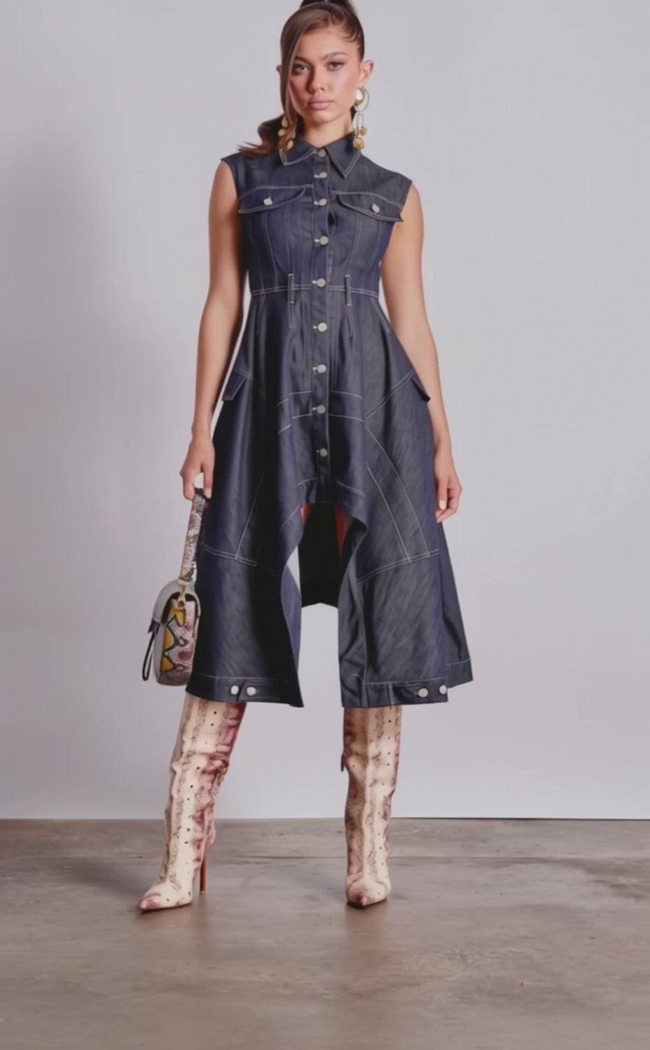 A video of a woman in the MADELINE BUTTON UP DENIM MIDI DRESS, featuring a sleeveless design and asymmetrical hem, styled with statement earrings, a handbag, and knee-high snakeskin boots.