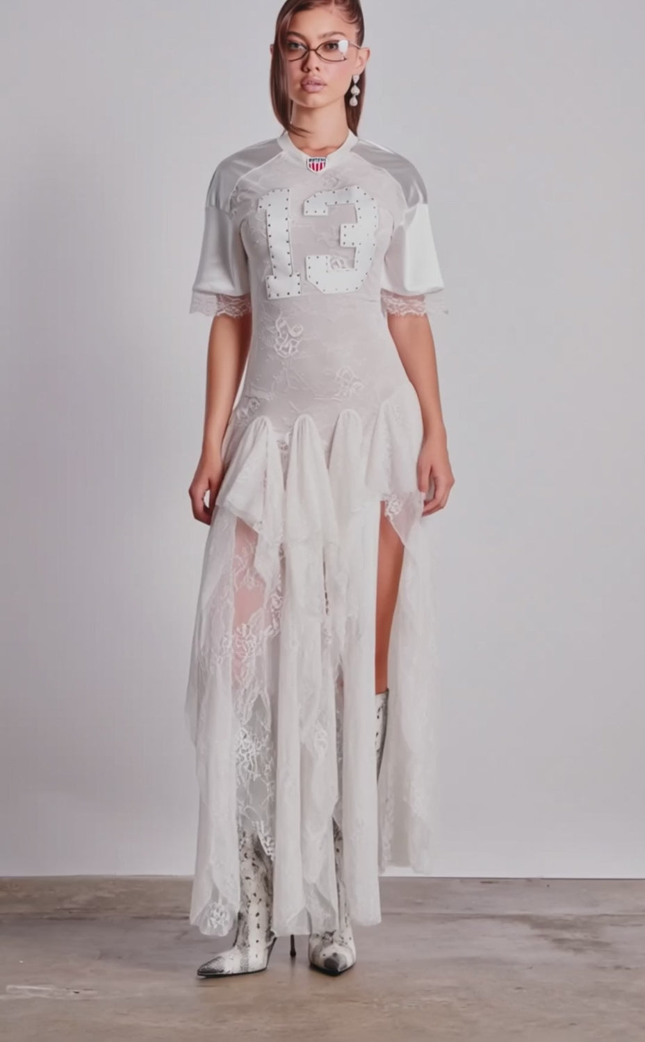 A video of a model wearing the KICK OFF LACE JERSEY MAXI DRESS IN WHITE with a high slit, clear glasses, and knee-high patterned boots.