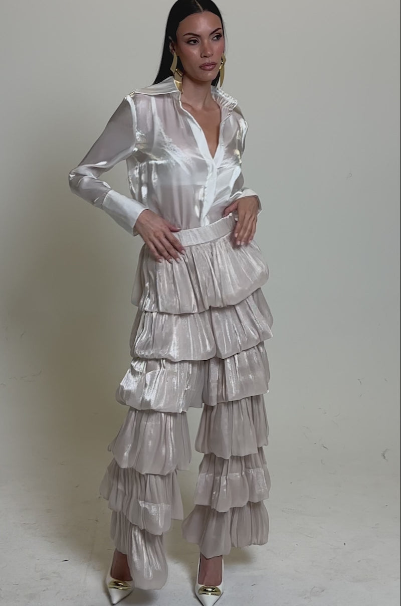 A video of a woman wearing a shiny white button-up blouse with long sleeves and the STAR OF THE NIGHT FLOUNCY PANT IN IVORY. She stands in high heels against a plain light background, looking confidently at the camera.