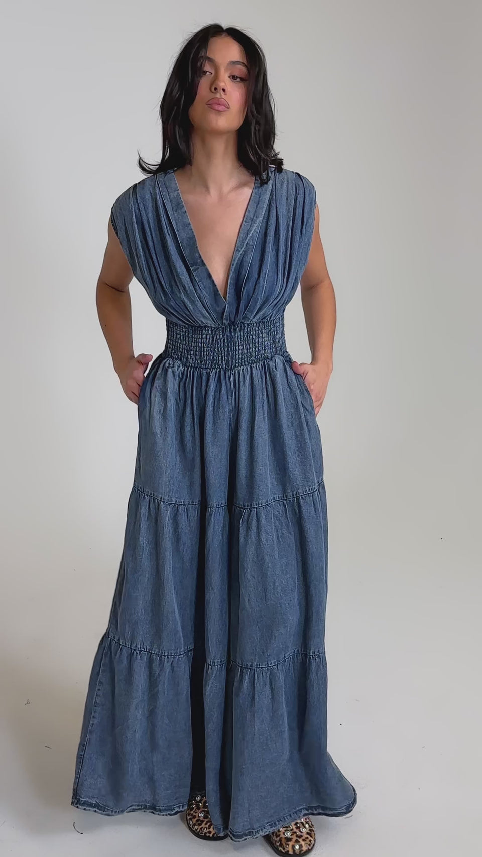 A video of a woman in the KELLY DENIM JUMPSUIT—a sleeveless, deep V-neck, floor-length blue denim style with a smocked waist and tiered skirt—paired with leopard print shoes.