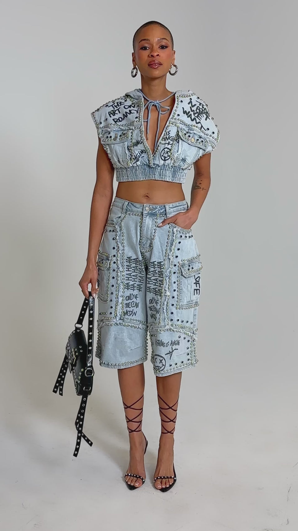 Video of woman modeling the GET INTO THE RHYTHM CROP HOODIE with high-waisted pants, metal studs, and graffiti graphics, she completes her street-chic look with hoop earrings and confidence.