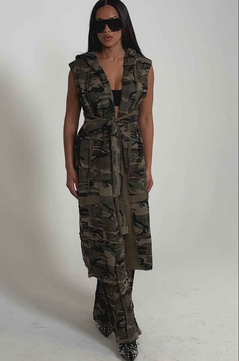 A video of a woman wearing the CAN'T SEE ME HOODED LONGLINE CAMO SWEATSHIRT, styled with black shoes, sunglasses, and bold accessories.