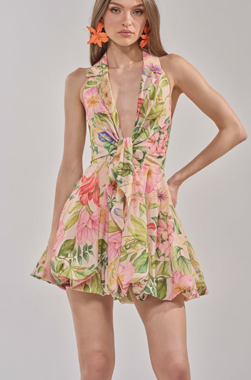 Video of woman modeling the SOFT LAUNCH FLORAL MINI DRESS IN BEIGE, featuring a sleeveless design, deep V-neckline, and flared skirt. She accessorizes with orange flower earrings for a chic vacation look.