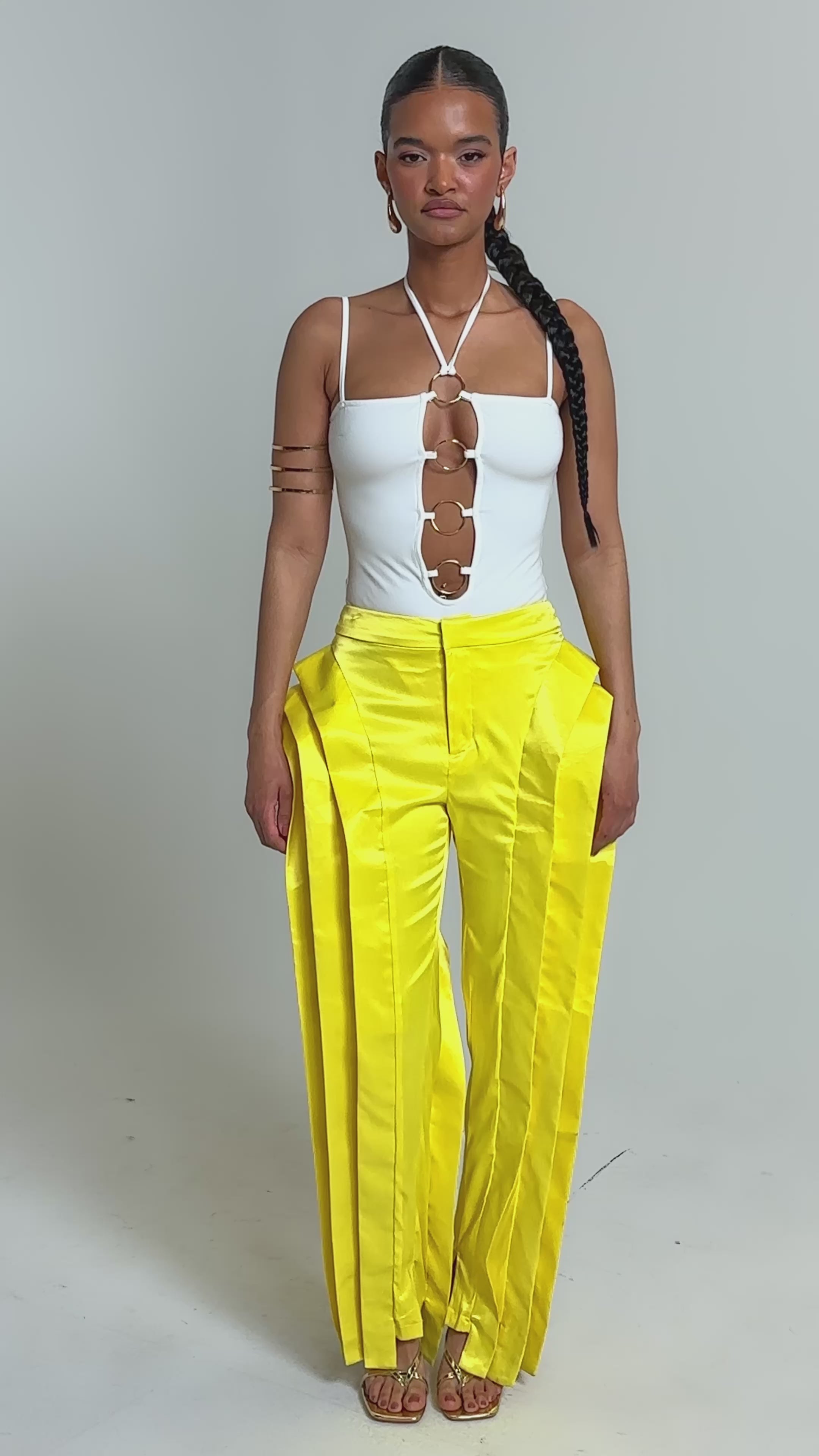 Video of woman modeling the FLY ME OUT BODYSUIT featuring a white halter neckline and gold ring hardware, paired with bold gold jewelry and yellow pleated pants, standing confidently against a plain background.