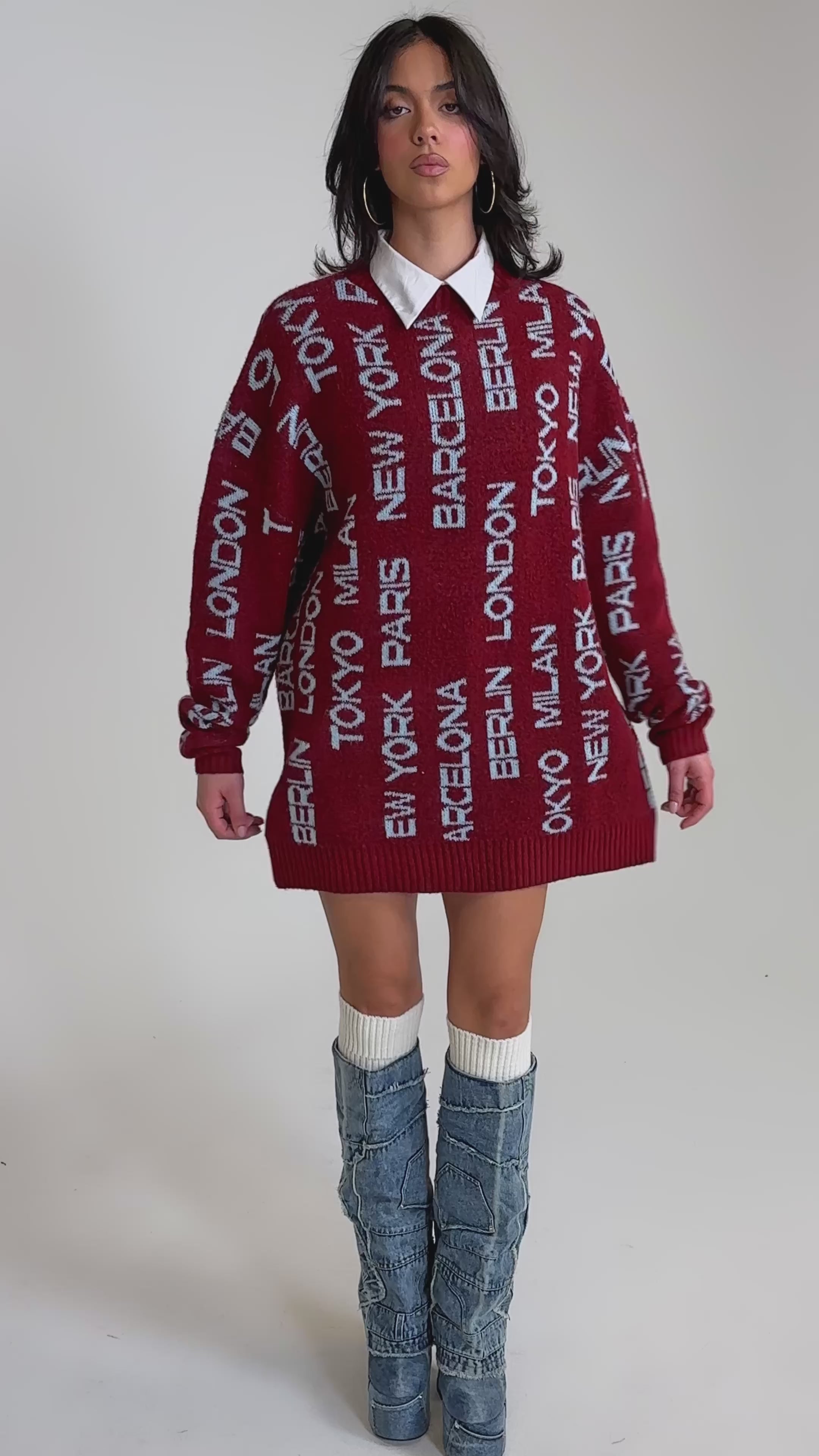 A video of a woman wearing the AROUND THE WORLD SWEATER MINI DRESS IN RED, featuring white city names, layered over a white collared shirt with tall slouchy denim platform boots. 