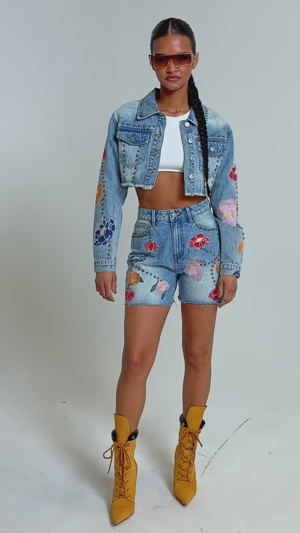 Video of woman modeling the LOTTIE EMBROIDERED DENIM BERMUDA SHORTS with floral appliqués, a cropped denim jacket, white crop top, yellow lace-up ankle boots, and sunglasses against a plain background.