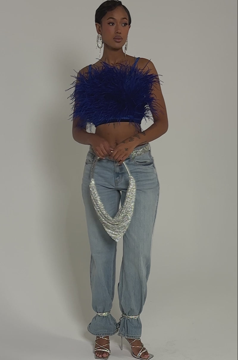 Video of woman modeling the BRIGHT LIGHTS FEATHER TOP—royal blue, velvet, cropped—paired with light blue jeans, sparkling earrings, and a green glittery purse. Her short hair is styled perfectly, with tattoos visible on her hands.