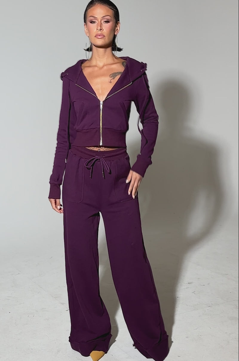 A video of a woman posing confidently in the SHE'S GONE, YOU'RE FINE WIDE LEG JOGGER—a deep purple matching cotton tracksuit with wide leg joggers and a zip-up hoodie. She pairs it with hoop earrings and slicked-back hair against a plain light background.