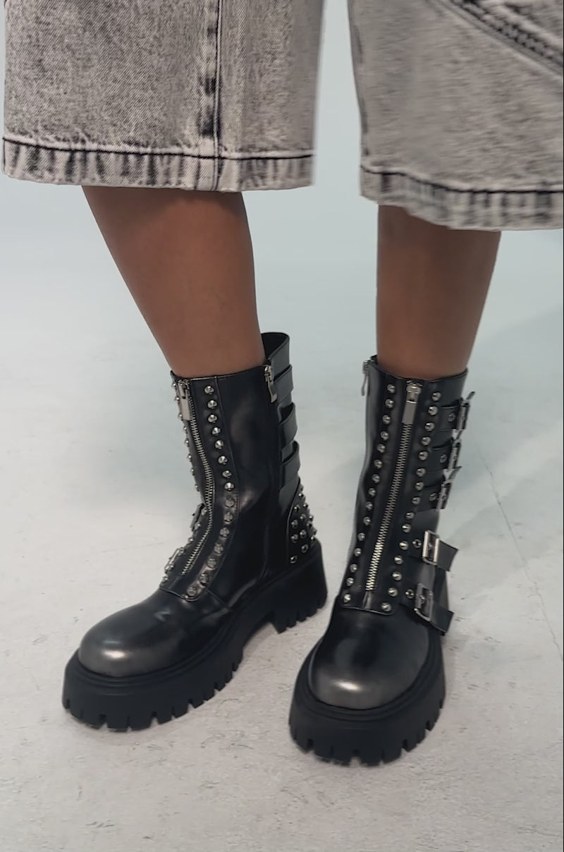 A video of a model wearing the AKIRA GOLD ESTERA CHARCOAL EMBELLISHED COMBAT BOOTIE which features black faux leather, four silver buckle straps, a side zipper, silver heel studs, and a chunky treaded sole.