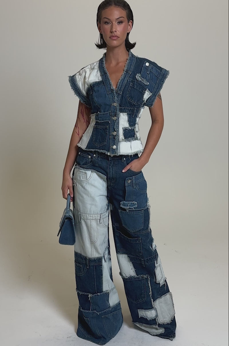 A video of a woman in the TOO MUCH GOING ON WIDE LEG DENIM, high waisted patchwork pants paired with a sleeveless button-up top and matching denim handbag, all featuring blue and white patchwork details.