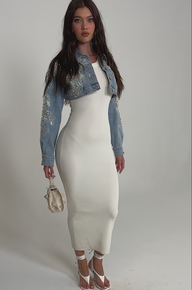 A video of a woman wearing a long white fitted dress, the BILLIONAIRE BELLE EMBELLISHED DENIM JACKET with floral embroidery, white strappy heels, and holding a small beige handbag.