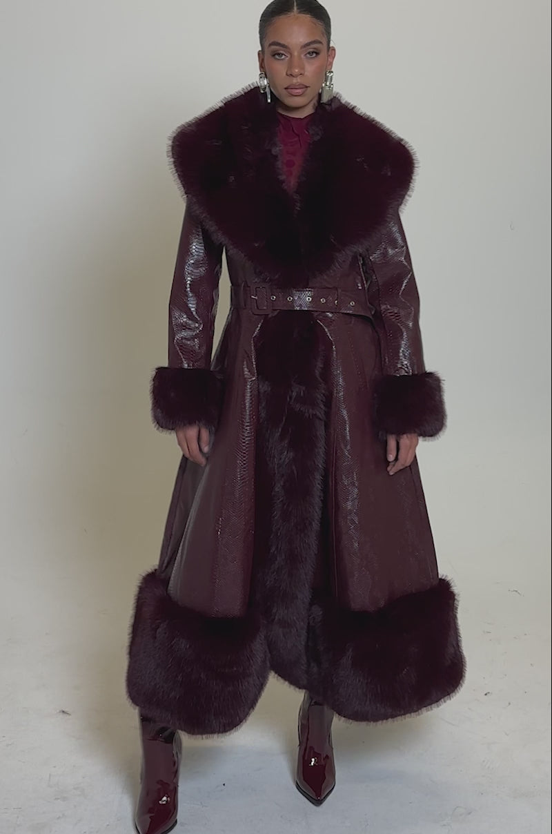 A video of a woman in the VIVIAN FUR TRIM TRENCH IN BURGUNDY, featuring a wide fur collar, fur cuffs, and hem. The belted waist flatters her figure as she pairs it with glossy burgundy boots for a polished look.